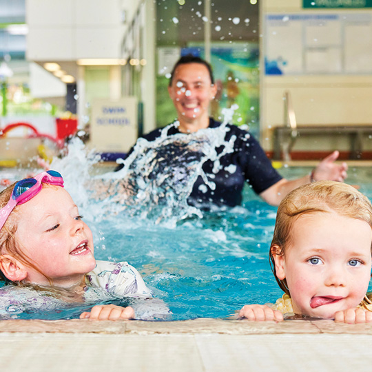 Children in water holding the edge of a pool with learn to swim instructor splashing in the background